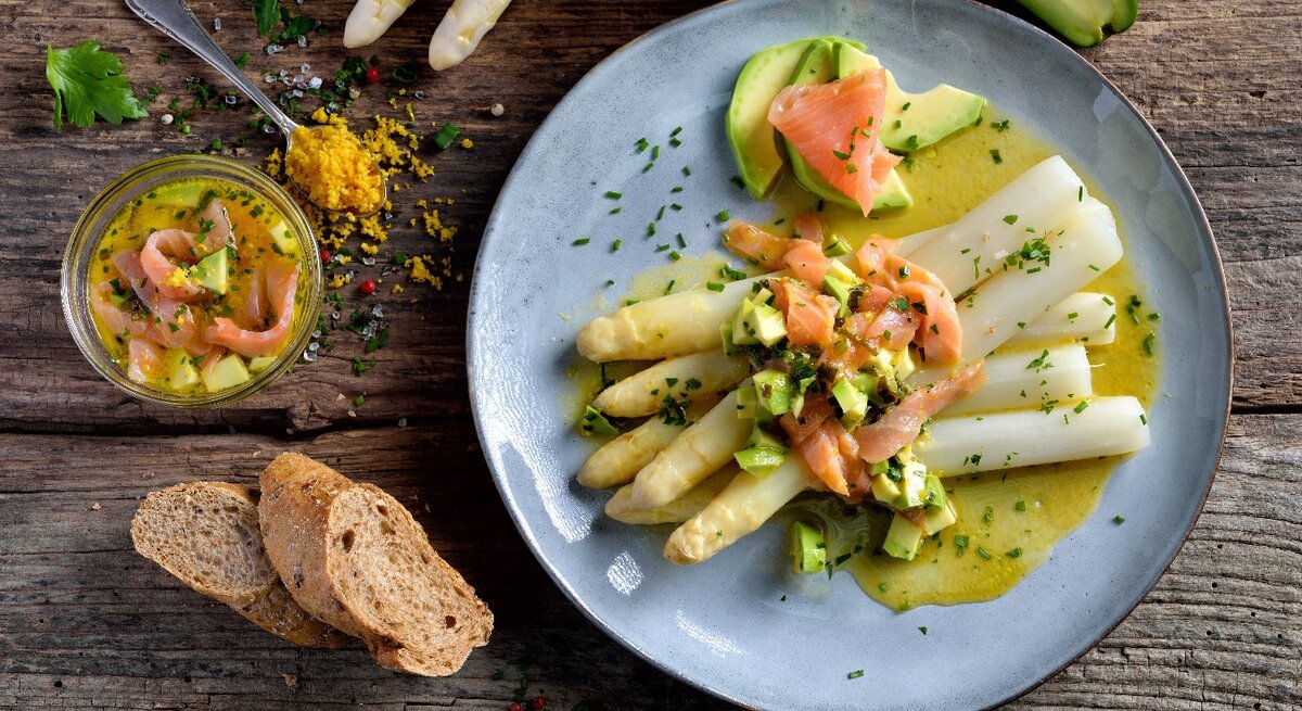Ein hellblauer Teller mit Spargel, Avocado, Räucherlachs und Zitronenbutter auf einem dunklen Holztisch. Daneben ein Teelöffel mit geriebenem Eigelb, der Zitronenbutter und rohen weißen Spargelstangen.