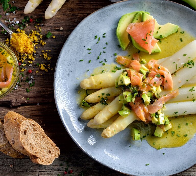 Ein hellblauer Teller mit Spargel, Avocado, Räucherlachs und Zitronenbutter auf einem dunklen Holztisch. Daneben ein Teelöffel mit geriebenem Eigelb, der Zitronenbutter und rohen weißen Spargelstangen. | © Shutterstock/ Karl Allgaeuer