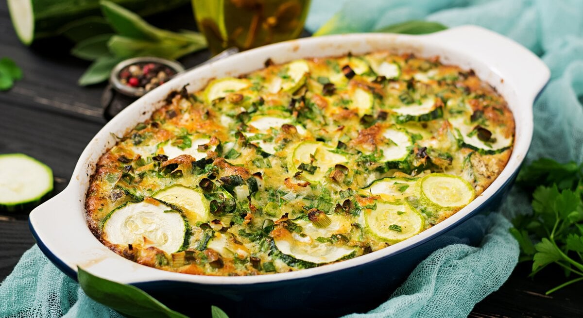 Kartoffel-Lauch-Gratin mit Zucchini in dunkelblauer Auflaufform, angerichtet auf einem blauen Tuch auf dunklem Holz. Umgeben ist die Speise von den Zutaten. 