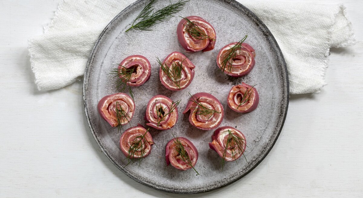 Rote-Bete-Pfannkuchenröllchen mit Lachs und Meerrettich, gerollt und in kleine Happen geschnitten serviert auf einem grauen Tisch auf einem weißen Tisch.