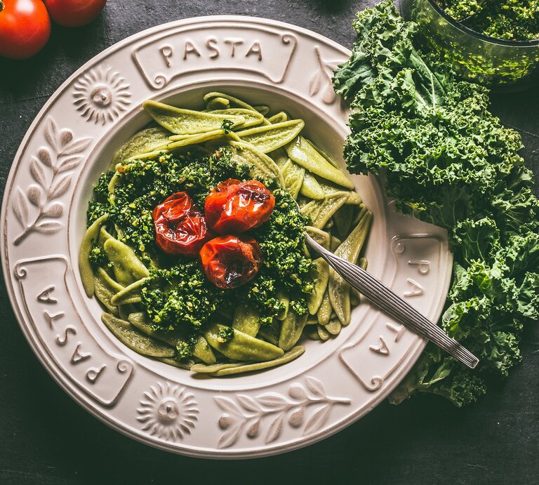 Ein Teller Grünkohl-Pasta mit Zitrone und Tomaten in der Draufsicht, daneben frischer Grünkohl und ganze Tomaten. | © Adobe Stock/ VICUSCHKA