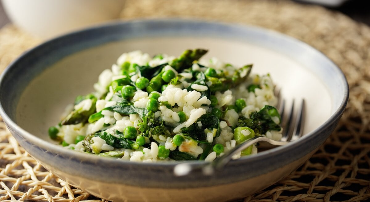 Ein Teller veganes Spargelrisotto mit Erbsen und Spinat, darin liegt eine Gabel.