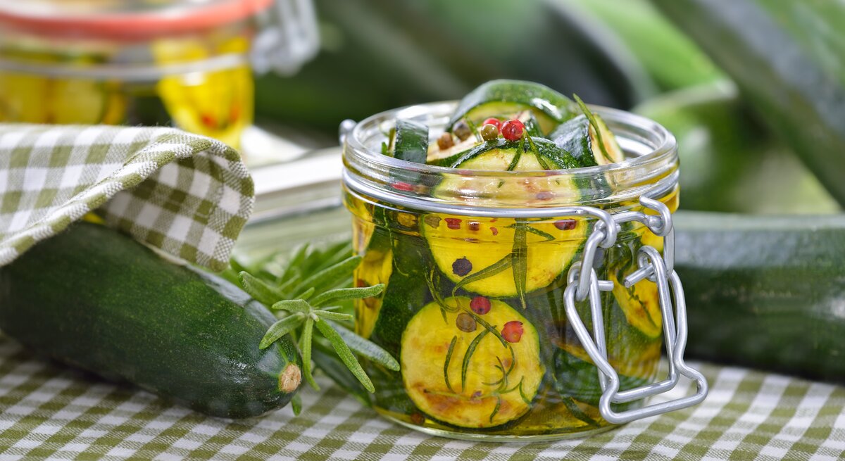 Zwei Weckgläser mit eingelegter Zucchini auf einer weiß-grün karierten Tischdecke mit Kräutern.