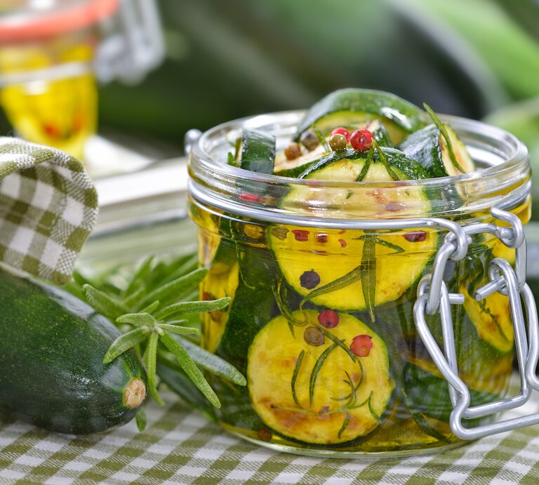 Zwei Weckgläser mit eingelegter Zucchini auf einer weiß-grün karierten Tischdecke mit Kräutern. | © Shutterstock/ Karl Allgaeuer