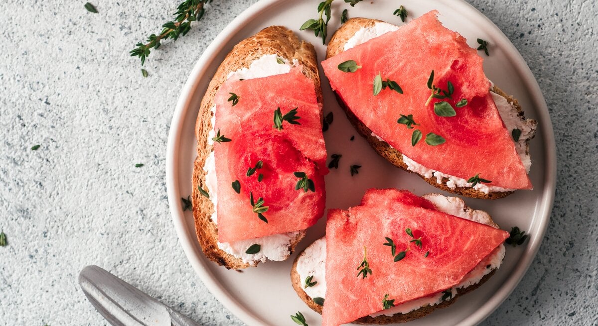Ein Teller mit Brotscheiben, auf denen Frischkäse und veganer Thunfisch aus Wassermelone liegt. Bestreut mit frischen Kräutern. Draufsicht.