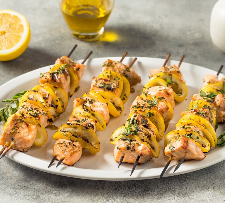 Gegrillte Lachsspieße mit Zucchini und Kräutermarinade auf weißem Teller auf grauem Untergrund. Daneben Kräuter, Zitrone und Olivenöl. | © Getty Images / bhofack2