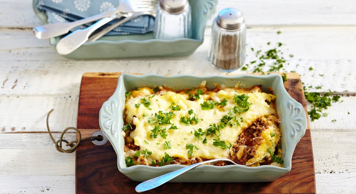 Eine Auflaufform mit Polenta-Hackfleischgratin. Daneben weiteres Besteck und Geschirr und Salz und Pfeffer. Das Ganze auf einem hölzernen Untergrund. 