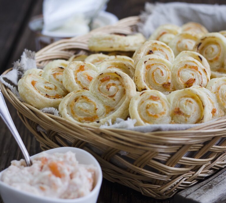 Blätterteigschnecken mit Lachs und Meerettich-Frischkäse, gestapelt in einem großen Korb, serviert mit der Lachs-Creme auf einem hölzernen Tisch.  | © StockFood / Tamara Staab