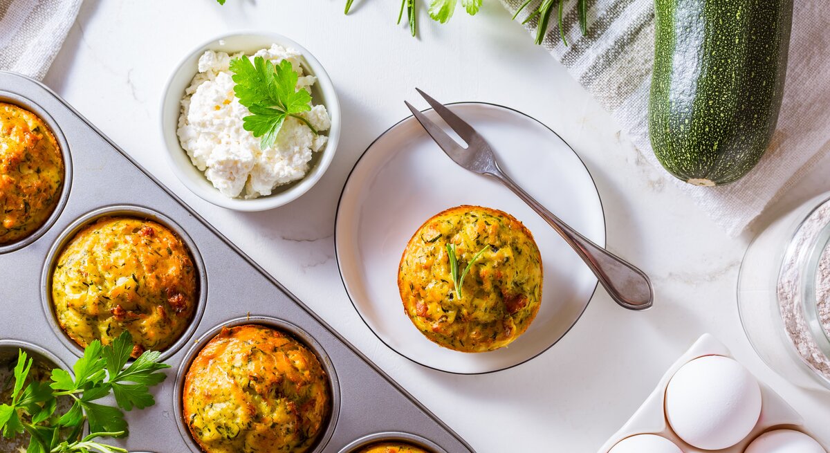 Die Muffin-Form mit den Muffins auf einem Tisch mit weißer Tischdecke. Daneben weiße Eier und ein Teller mit einem Muffin aus der Form.