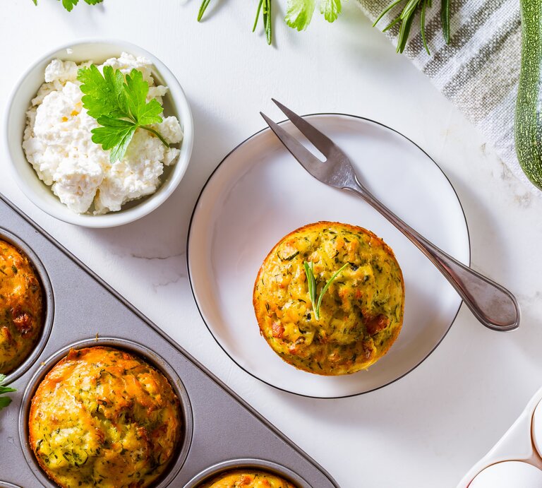 Die Muffin-Form mit den Muffins auf einem Tisch mit weißer Tischdecke. Daneben weiße Eier und ein Teller mit einem Muffin aus der Form. | © AdboeStock/ Brebca