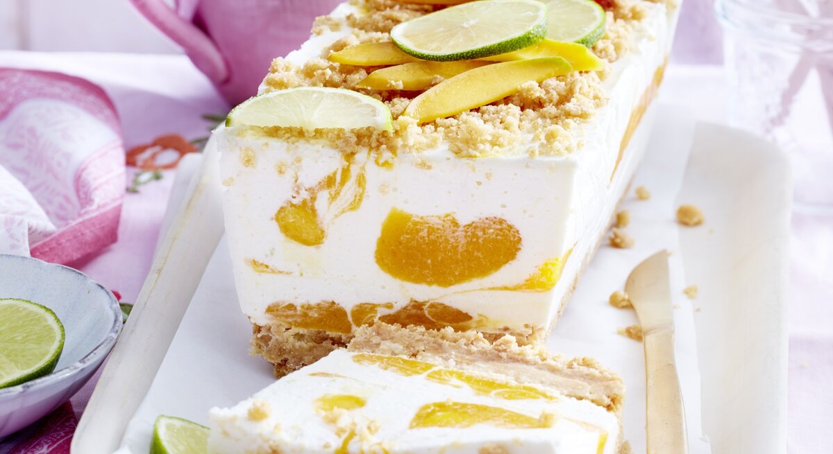 Mango-Kokos-Cheesecake in Kastenform, garniert mit Streuseln und frischen Mango und Limetten-Stückchen und serviert auf einer weißen Form auf einem rosanen Tisch-Deck. 
