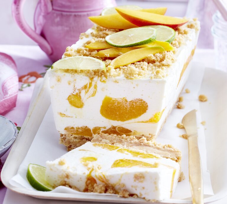 Mango-Kokos-Cheesecake in Kastenform, garniert mit Streuseln und frischen Mango und Limetten-Stückchen und serviert auf einer weißen Form auf einem rosanen Tisch-Deck.  | © EAT CLUB / Isabell Triemer