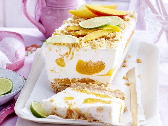 Mango-Kokos-Cheesecake in Kastenform, garniert mit Streuseln und frischen Mango und Limetten-Stückchen und serviert auf einer weißen Form auf einem rosanen Tisch-Deck.  | © EAT CLUB / Isabell Triemer