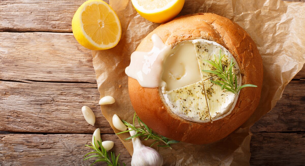 Auf einem hölzernem Untergrund liegt ein Packpapier. Darauf liegt ein ausgehöhlter Brotlaib. In dem Brotlaib ist ein Camembert, auf dem ein Zweig Rosmarin zu sehen. Neben dem Brot liegen eine Knoblauchknolle, Knoblauchzehen und ein weiterer Rosmarinzweig.