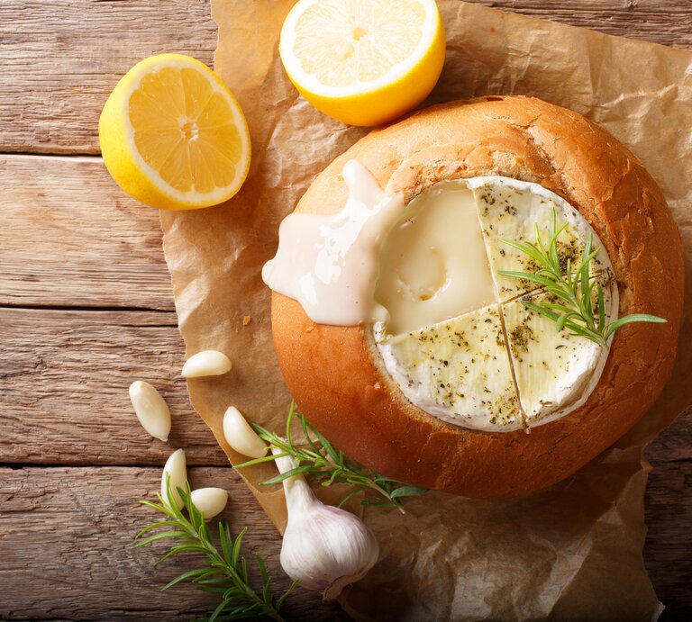 Auf einem hölzernem Untergrund liegt ein Packpapier. Darauf liegt ein ausgehöhlter Brotlaib. In dem Brotlaib ist ein Camembert, auf dem ein Zweig Rosmarin zu sehen. Neben dem Brot liegen eine Knoblauchknolle, Knoblauchzehen und ein weiterer Rosmarinzweig. | © Getty Images / ALLEKO