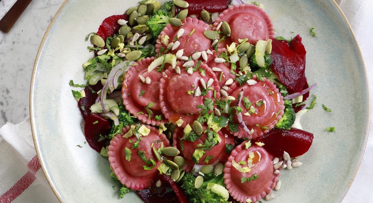 Salat mit Rote-Bete-Tortellini, Zwiebeln, frischer Rote Bete, Brokkoli, Kernen und Kräutern, serviert in einer hellen Schale auf einem Marmortisch, von oben fotografiert. 