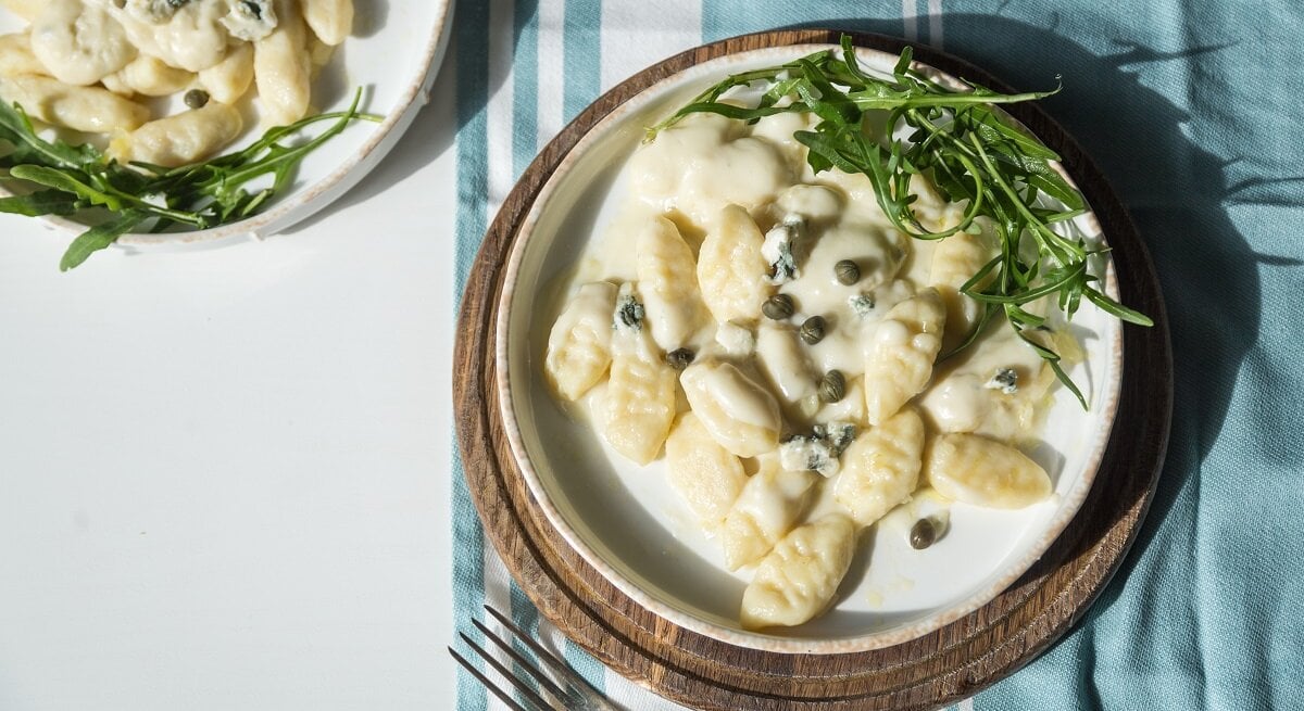 Zwei Teller Gnocchi mit Gorgonzola-Kapern-Soße stehen auf einem Tisch. Ein Teller ist nur halb zu sehen. Der andere ganz. Letzterer steht auf einem gestreiften Tuch. Auch die Spitzen einer Gabel sind auf dem Bild.