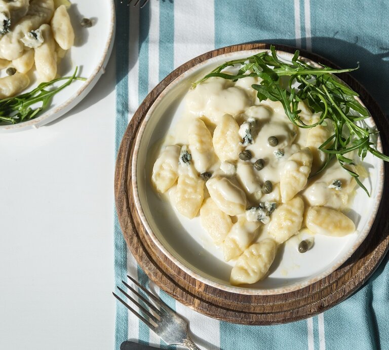 Zwei Teller Gnocchi mit Gorgonzola-Kapern-Soße stehen auf einem Tisch. Ein Teller ist nur halb zu sehen. Der andere ganz. Letzterer steht auf einem gestreiften Tuch. Auch die Spitzen einer Gabel sind auf dem Bild. | © Shutterstock / Sofiia Popovych