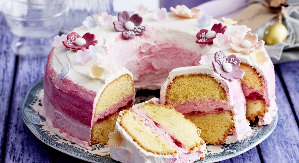 Auf einem Teller liegt ein Blütenkranz-Kuchen. Er ist mit Sahne eingestrichen und mit essbaren Blüten dekoriert. Man erkennt die Erbdeerfüllung in der Mitte.