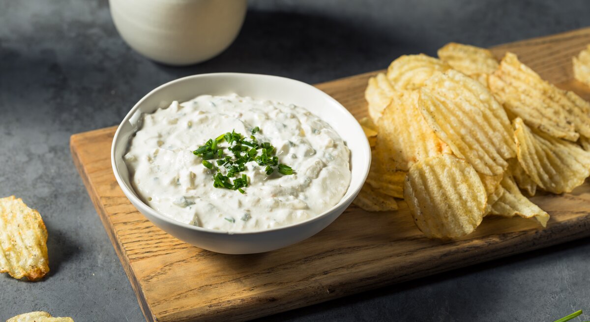 Französischer Zwiebeldip in weißer Schale auf Holzbrett auf dunkelgrauem Tisch. Auf dem Brett neben dem Dipschälchen geriffelte Chips. Am oberen Bildrand ein Kännchen mit Sahne. Gräser und Chips als Deko in Bild. Frontalansicht.