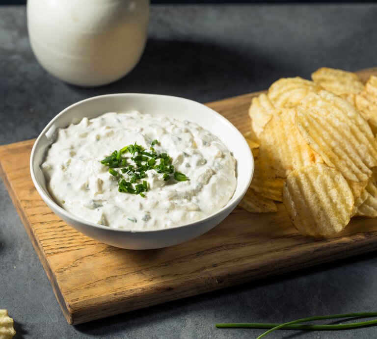 Französischer Zwiebeldip in weißer Schale auf Holzbrett auf dunkelgrauem Tisch. Auf dem Brett neben dem Dipschälchen geriffelte Chips. Am oberen Bildrand ein Kännchen mit Sahne. Gräser und Chips als Deko in Bild. Frontalansicht. | © Getty Images/bhofack2