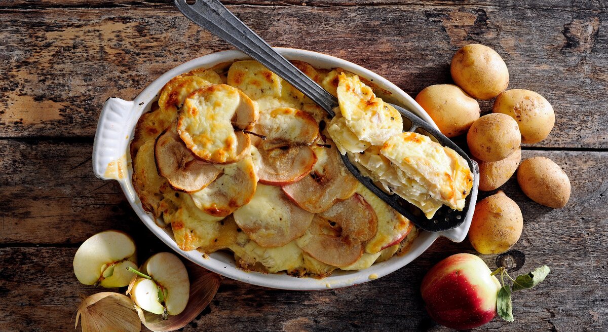 Mit Käse überbackener Kartoffel-Apfel-Auflauf in weißer Auflaufform auf hölzernem Untergrund. Darum herum sind Kartoffeln und Äpfel verteilt. 