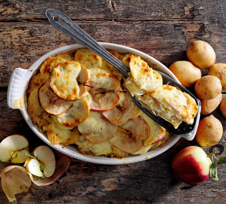Mit Käse überbackener Kartoffel-Apfel-Auflauf in weißer Auflaufform auf hölzernem Untergrund. Darum herum sind Kartoffeln und Äpfel verteilt.  | © Shutterstock/zebratomato