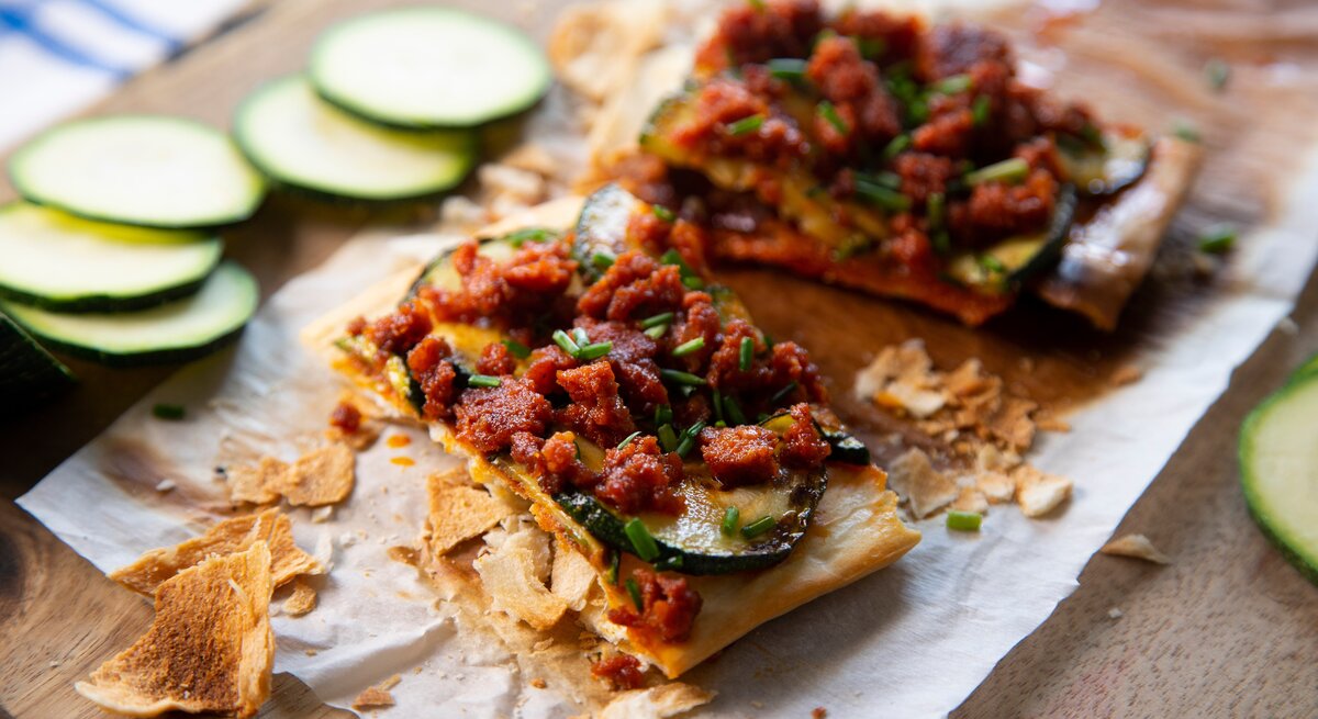 Spanische Maismehlpizza Coca Mallorcina auf Backpapier und Holzbrett. Daneben Zucchinischeiben. Draufsicht.
