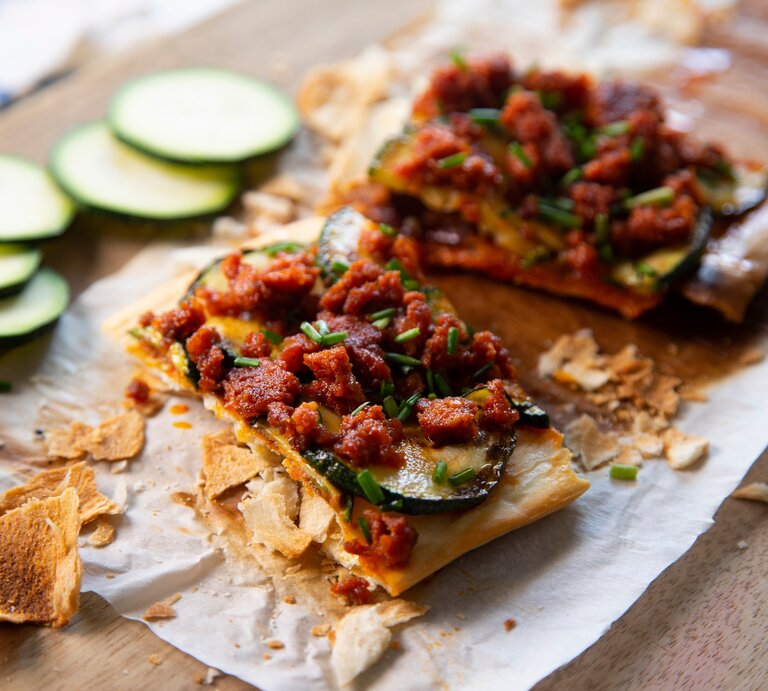 Spanische Maismehlpizza Coca Mallorcina auf Backpapier und Holzbrett. Daneben Zucchinischeiben. Draufsicht. | © shutterstock / Lecker Studio