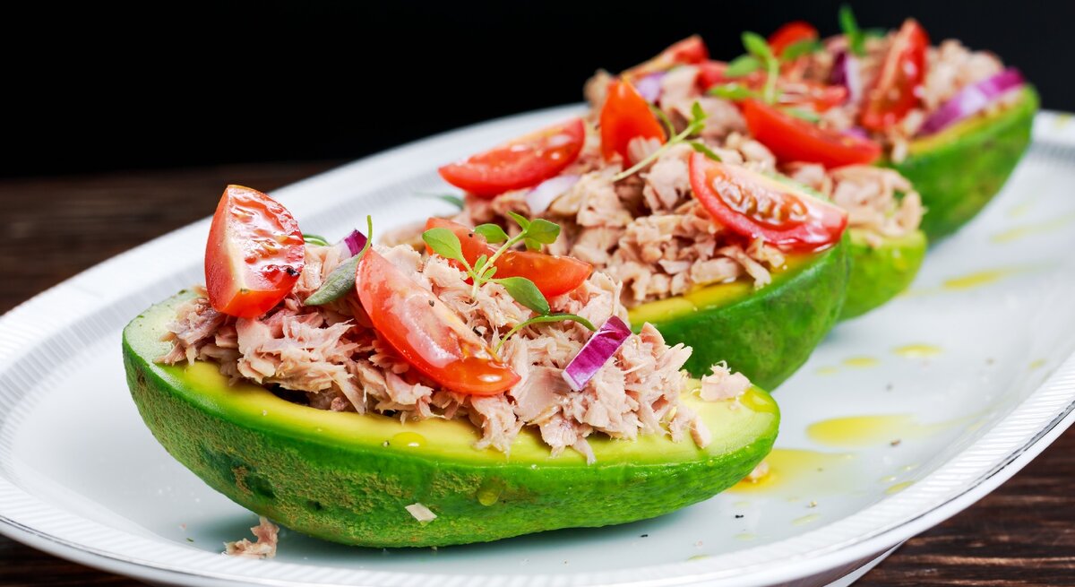Ein weißer länglicher Teller mit vier Avocadohälften, in denen der Thunfischsalat angerichtet ist vor schwarzem Hintergrund.