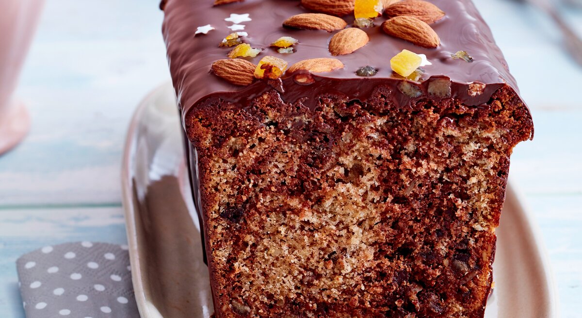 Angeschnittener Marmor-Gewürzkuchen mit Schokoladen-Glasur, Mandeln, Zuckersternen und Orangeat auf rosafarbenem Teller, weiß gepunkteter grauer Serviette und hellblauem Tischtuch in der Frontalsicht.