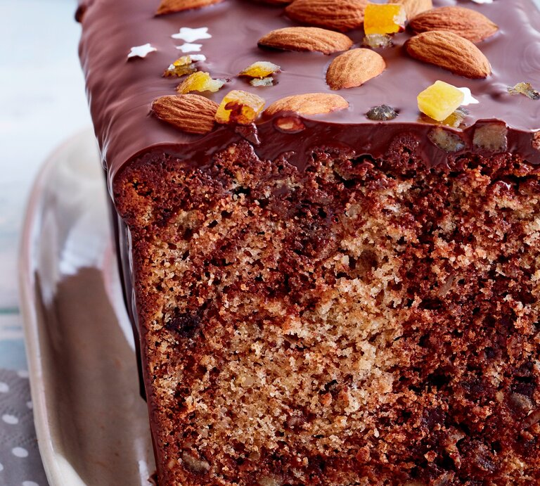 Angeschnittener Marmor-Gewürzkuchen mit Schokoladen-Glasur, Mandeln, Zuckersternen und Orangeat auf rosafarbenem Teller, weiß gepunkteter grauer Serviette und hellblauem Tischtuch in der Frontalsicht. | © EATCLUB / Isabell Triemer