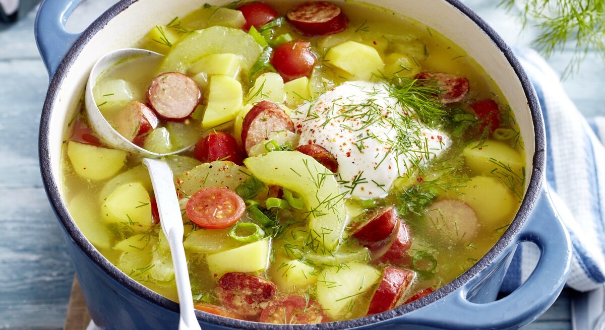 In einem Topf ist eine Portion Schmorgurken-Kabanossi-Eintopf angerichtet. Sie ist mit Dill und saurer Sauce serviert. 