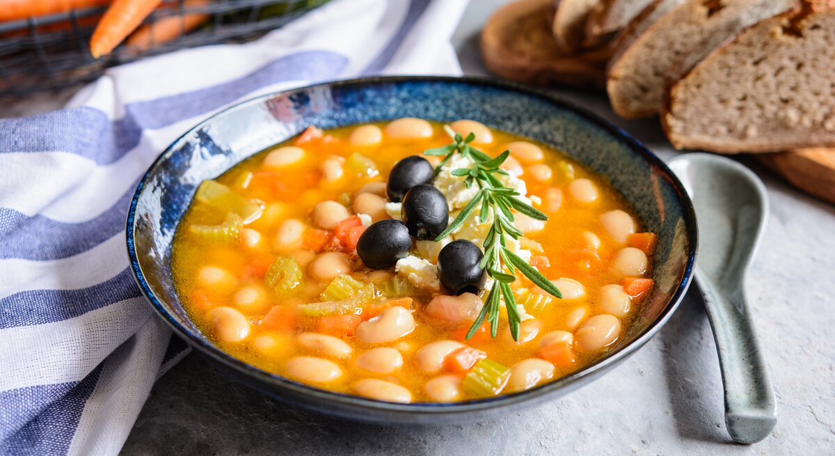 Bohnensuppe in einer blauen Schale auf grauem Tisch. Im Hintergrund ein Brotkorb, ein blau-weißes Tuch und ein Bund Karotten.