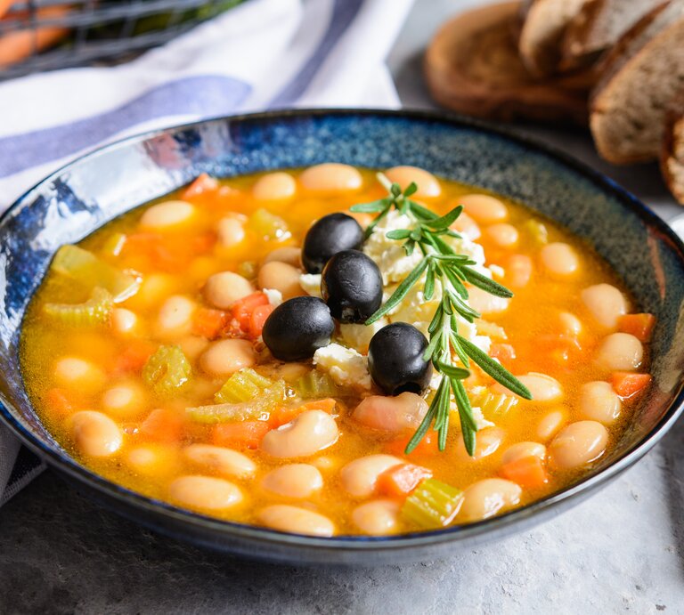 Bohnensuppe in einer blauen Schale auf grauem Tisch. Im Hintergrund ein Brotkorb, ein blau-weißes Tuch und ein Bund Karotten. | © Gettyimages/ NoirChocolate