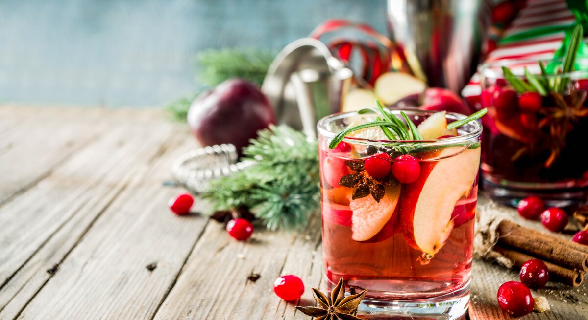 Ein Glas winterliche Sangria mit Apfel und Orange sowie Cranberrys, Anis und Rosmarin. Im Hintergrund sind unscharf die Zutaten zu erkennen.