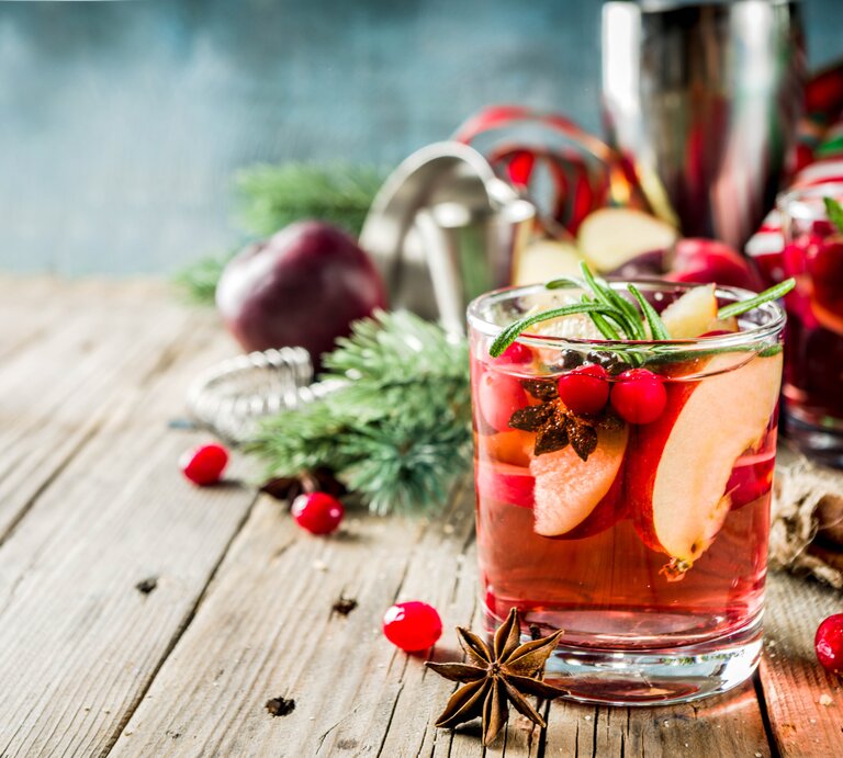 Ein Glas winterliche Sangria mit Apfel und Orange sowie Cranberrys, Anis und Rosmarin. Im Hintergrund sind unscharf die Zutaten zu erkennen. | © Getty Images/ Rimma_Bondarenko