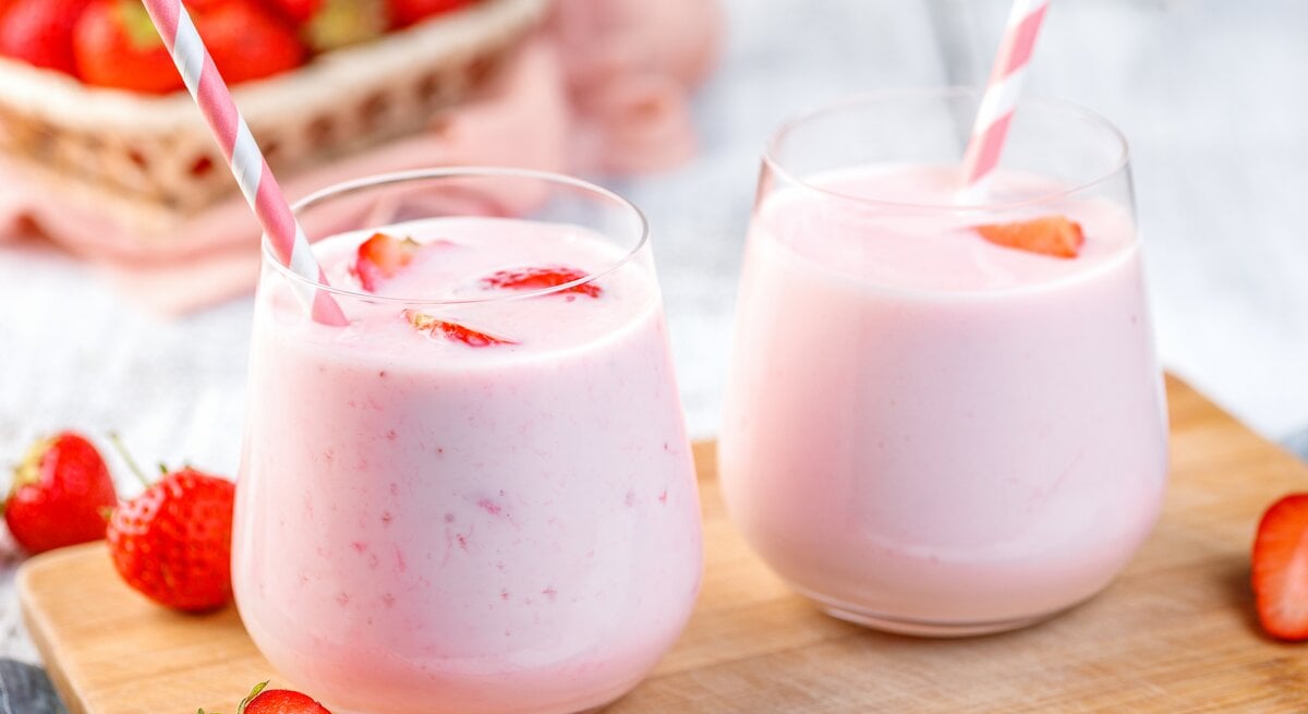 Zwei Gläser mit Erdbeermilchshake und gestreiftem Strohhalm stehen auf einem Holzbrett. Daneben liegen Erdbeeren. 