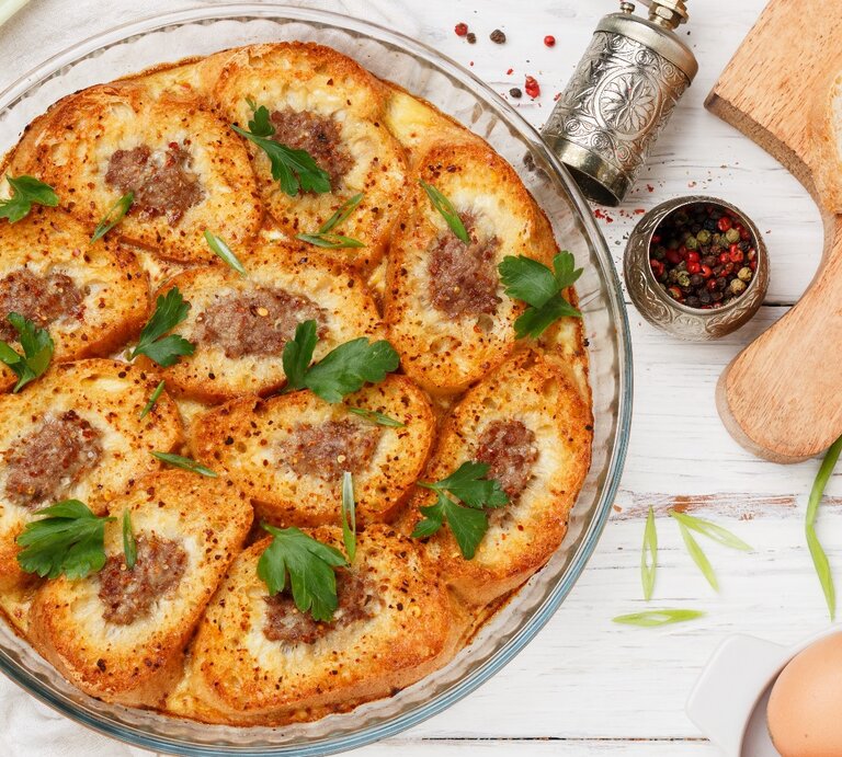 Eine Glasform mit den überbackenen Omelett-Baguettescheiben auf einem hellen Holztisch mit Eierschalen, Petersilie und geschnittenem Bagutte. | © Shutterstock/ Viktory Panchenko
