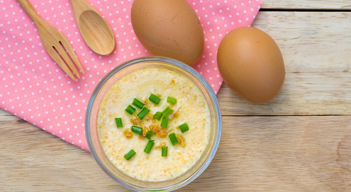 Eine Glasschüssel mit gedämpften Eierpudding mit frischen Frühlingszwiebeln auf einem roten Küchentuch neben Holzbesteck und Eiern von oben.