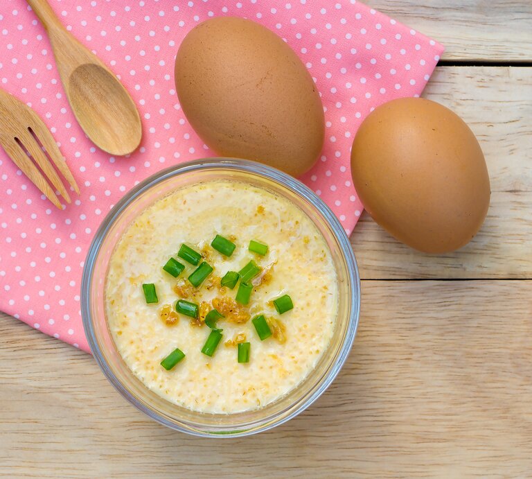 Eine Glasschüssel mit gedämpften Eierpudding mit frischen Frühlingszwiebeln auf einem roten Küchentuch neben Holzbesteck und Eiern von oben. | © Getty Images/ PitchyPix
