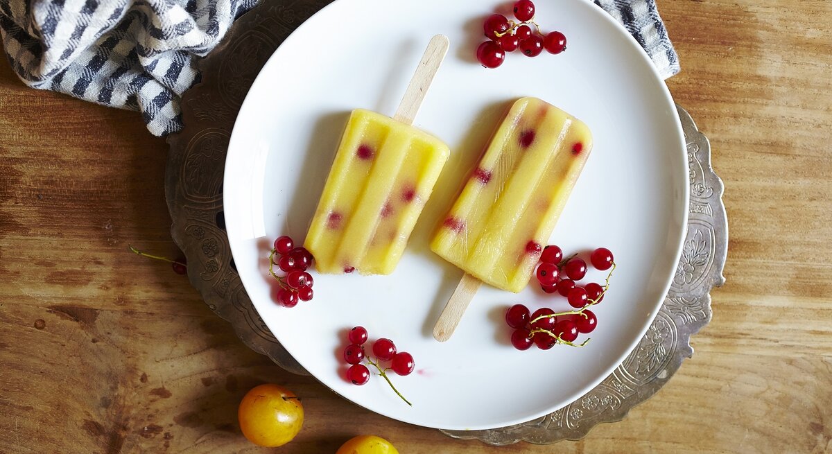 Zwei selbstgemachte Mirabellen-Preiselbeer Eis am Stiel auf einem weißen Teller mit frischen Preiselbeeren und Mirabellen umgeben, von oben vor einem Holzhintergrund.
