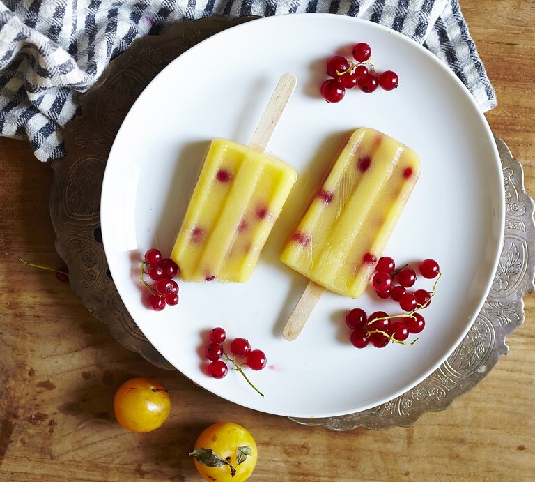 Zwei selbstgemachte Mirabellen-Preiselbeer Eis am Stiel auf einem weißen Teller mit frischen Preiselbeeren und Mirabellen umgeben, von oben vor einem Holzhintergrund. | © Getty Images /Cavan Images