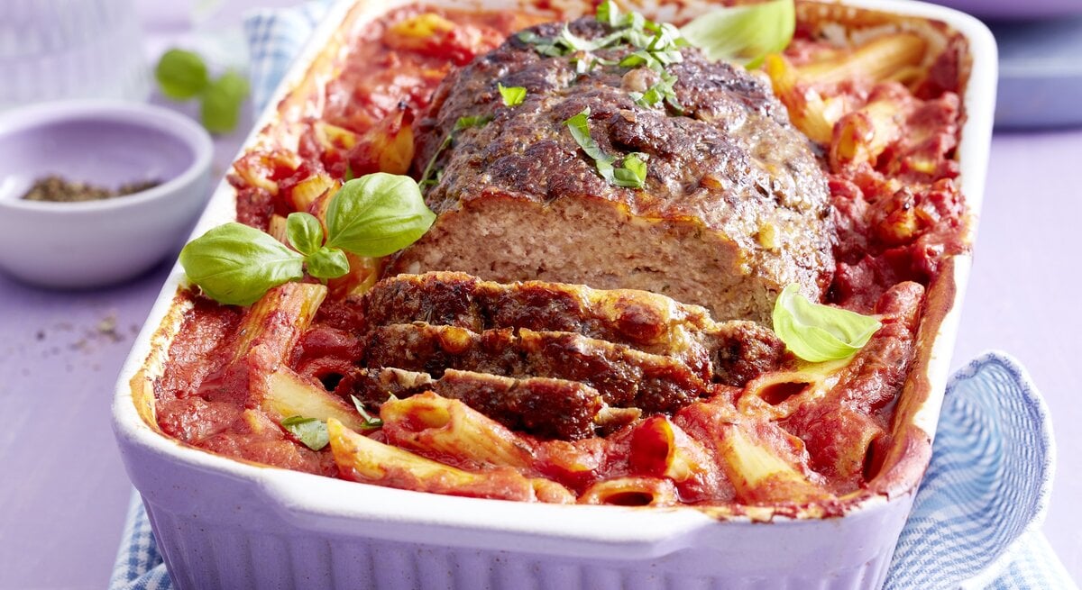 Ein One-Pot-Braten mit Nudeln liegt in einer weißen Auflaufform. Der Braten ist angerichtet mit Basilikum. Die Form steht auf einem blau weißen Tuch. Im Hintergrund steht eine Schale mit Pfeffer und verschiedenes Geschirr.