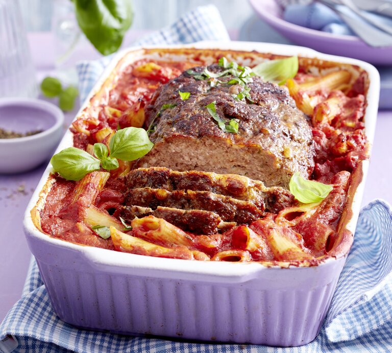 Ein One-Pot-Braten mit Nudeln liegt in einer weißen Auflaufform. Der Braten ist angerichtet mit Basilikum. Die Form steht auf einem blau weißen Tuch. Im Hintergrund steht eine Schale mit Pfeffer und verschiedenes Geschirr. | © EAT CLUB/Isabell Triemer