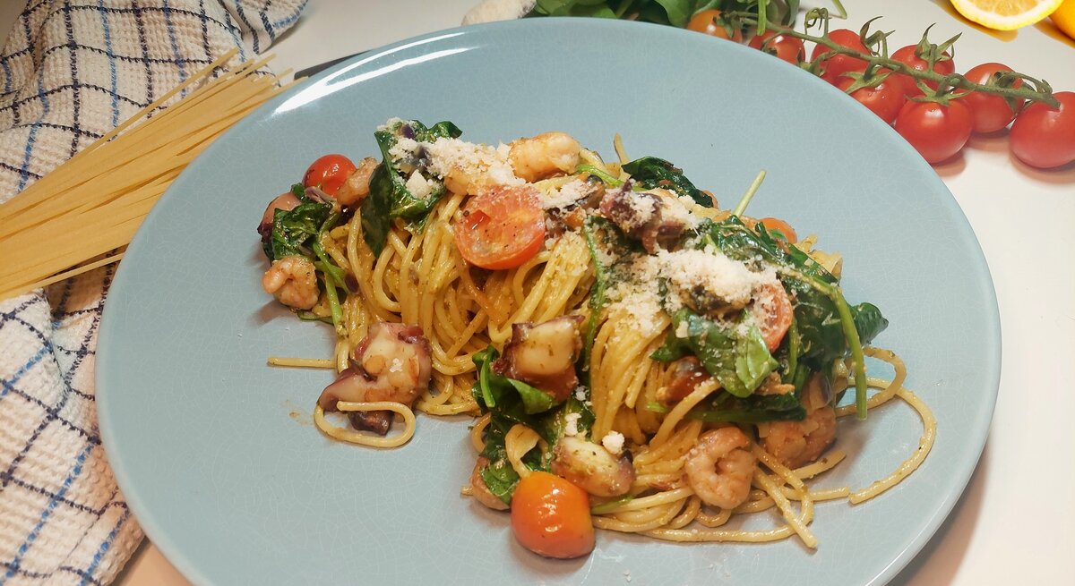 Blauer Teller mit den Zitronenspaghetti und Meersfrüchten. Im Hintergrund Nudeln Tomaten und Zitronen.