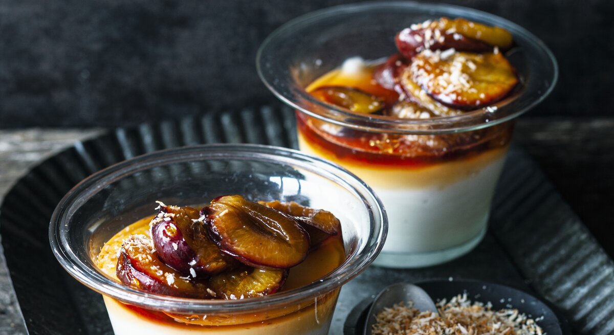 Zwei Portionen Kokospudding mit karamellisierten Zwetschgen stehen auf einem gemusterten schwarzen Teller auf einem Holztisch. Der Pudding ist angerichtet mit Kokosflocken.
