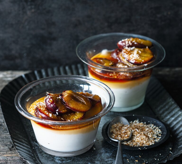 Zwei Portionen Kokospudding mit karamellisierten Zwetschgen stehen auf einem gemusterten schwarzen Teller auf einem Holztisch. Der Pudding ist angerichtet mit Kokosflocken. | © Stockfood/Gräfe & Unzer Verlag / Schardt, Wolfgang
