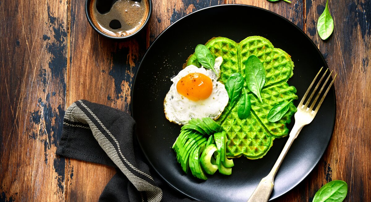 Aus der Vogelperspektive: Auf einem Holztisch liegt ein schwarzer Teller. Auf dem Teller liegt eine grüne Waffel. Auf der grünen Waffel ist ein gebratenes Ein. Daneben liegen Avocado-Scheiben sowie einige Spinatblätter.