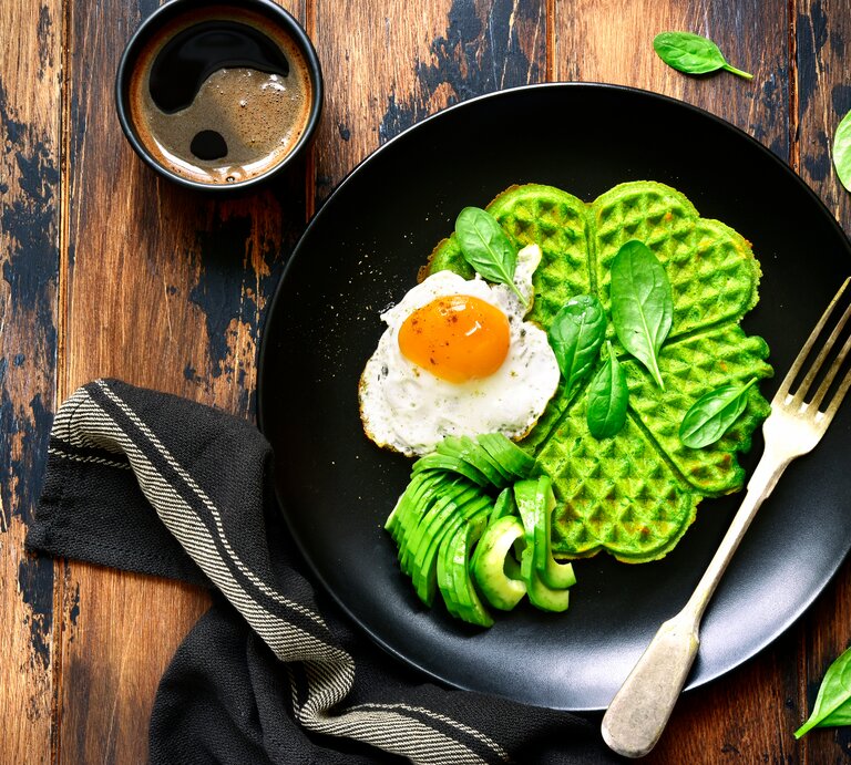 Aus der Vogelperspektive: Auf einem Holztisch liegt ein schwarzer Teller. Auf dem Teller liegt eine grüne Waffel. Auf der grünen Waffel ist ein gebratenes Ein. Daneben liegen Avocado-Scheiben sowie einige Spinatblätter. | © Shutterstock / Liliya Kandrashevich